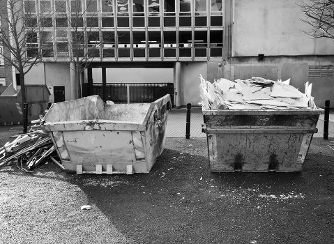 Two large industrial wheelie bins are positioned side by side on a paved area in an outdoor urban setting, with a modern building in the background. The left bin, made of metal with a weathered, slightly rusted finish, contains broken pieces of concrete, masonry, and debris, some protruding over its edges. The right bin, also metal but darker in appearance, is filled with crumpled and torn paper, cardboard, and other miscellaneous waste, piled unevenly. Surrounding the waste bins is a small amount of scattered rubble and dust on the ground, with a few metal supports or bars lying nearby on the left. The scene is lit by natural daylight, with the building's large windowed façade visible behind, indicating an urban environment that could be associated with house clearance or waste disposal services like those offered by House Clearance Thamesmead. The image emphasizes the practical aspect of rubbish removal services involving bulky waste collection and disposal within the Thamesmead area, inferring a typical street scene for waste management outside residential or commercial premises in southeast London.