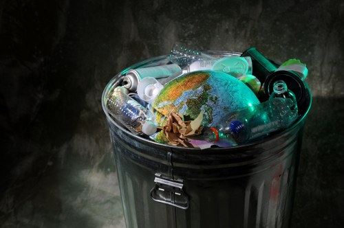 A metal rubbish bin with a secured lid, filled with various waste items including transparent plastic bottles, aluminum spray cans, and crumpled brown paper. The bin is positioned against a dark, textured surface, highlighting the reflective surfaces of the containers and the bright colors of the bottles and cans. The waste appears to be a mix of recyclable and non-recyclable materials, typical of household rubbish collected for disposal or recycling in Thamesmead. The environment suggests an indoor or covered outdoor setting, with natural or artificial lighting casting subtle reflections on the metallic surface of the bin. This image represents a typical waste collection scenario handled by rubbish removal services such as those provided by House Clearance Thamesmead, which manage household waste removal efficiently and professionally within the local area postcode and town.